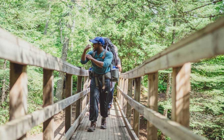 A person flexes their muscle as they walk across a wooden bridge toward the camera. There are dense green trees behind them.